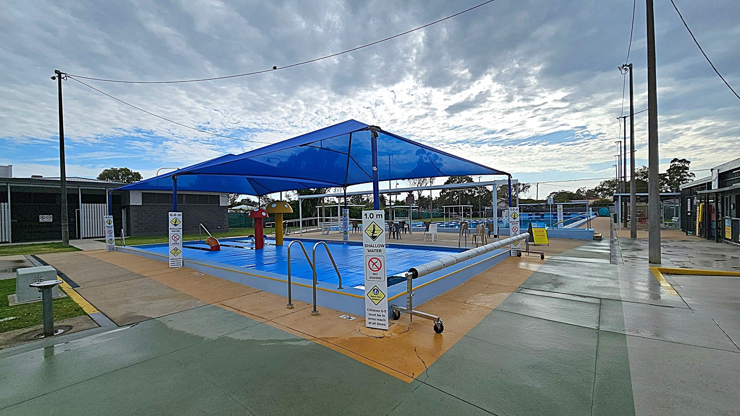 An outdoor swimming pool area with a blue shade canopy over a shallow pool. The pool is surrounded by a paved area with safety signs indicating water depth and no diving. On the left, there is a dark building with a white gate. On the right, another building with open entrances. Several chairs are scattered around the poolside. The ground is a mix of green and beige concrete, and tall light poles are visible around the area. Trees are in the background. An outdoor swimming pool area with a blue shade canopy over a shallow pool. The pool is surrounded by a paved area with safety signs indicating water depth and no diving. On the left, there is a dark building with a white gate. On the right, another building with open entrances. Several chairs are scattered around the poolside. The ground is a mix of green and beige concrete, and tall light poles are visible around the area. Trees are in the background.