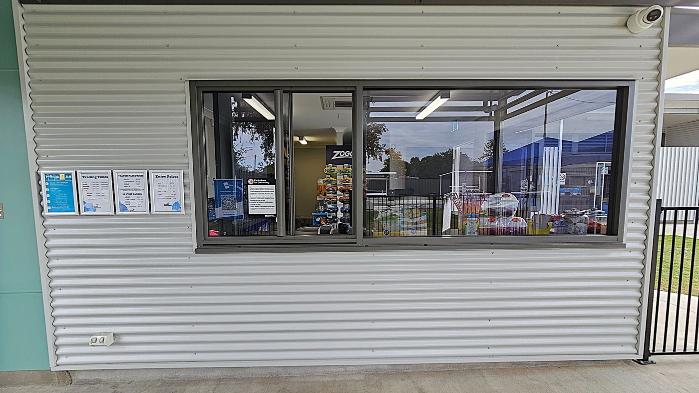 Entrance to the Oakey Aquatic Centre with a sign displaying the name and opening hours. The entrance is flanked by a corrugated metal building on the left and a black metal fence on the right. Beyond the entrance, there are blue shade structures over the pool area. A "No Smoking" sign is visible on the fence. The ground is paved with concrete, and there is a grassy area to the right. Trees are visible in the background. Entrance to the Oakey Aquatic Centre with a sign displaying the name and opening hours. The entrance is flanked by a corrugated metal building on the left and a black metal fence on the right. Beyond the entrance, there are blue shade structures over the pool area. A "No Smoking" sign is visible on the fence. The ground is paved with concrete, and there is a grassy area to the right. Trees are visible in the background.