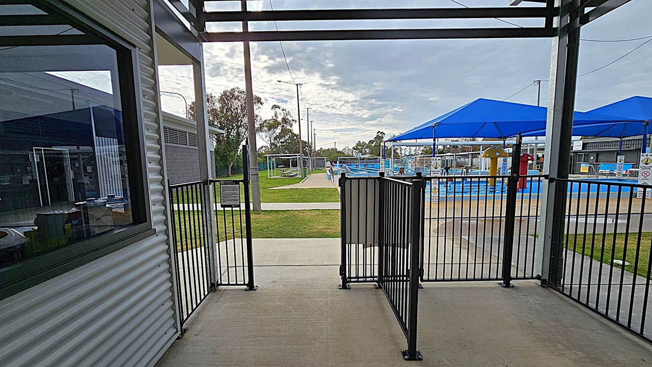 A view from a shaded area with a metal railing gate in the foreground. To the left, there's a corrugated metal building with a window. In the background, a grassy area leads to a swimming pool with blue shade structures. The pool area has colourful play equipment and is surrounded by a fence. Trees and utility poles are visible in the distance. The ground is a mix of concrete and grass. A view from a shaded area with a metal railing gate in the foreground. To the left, there's a corrugated metal building with a window. In the background, a grassy area leads to a swimming pool with blue shade structures. The pool area has colourful play equipment and is surrounded by a fence. Trees and utility poles are visible in the distance. The ground is a mix of concrete and grass.