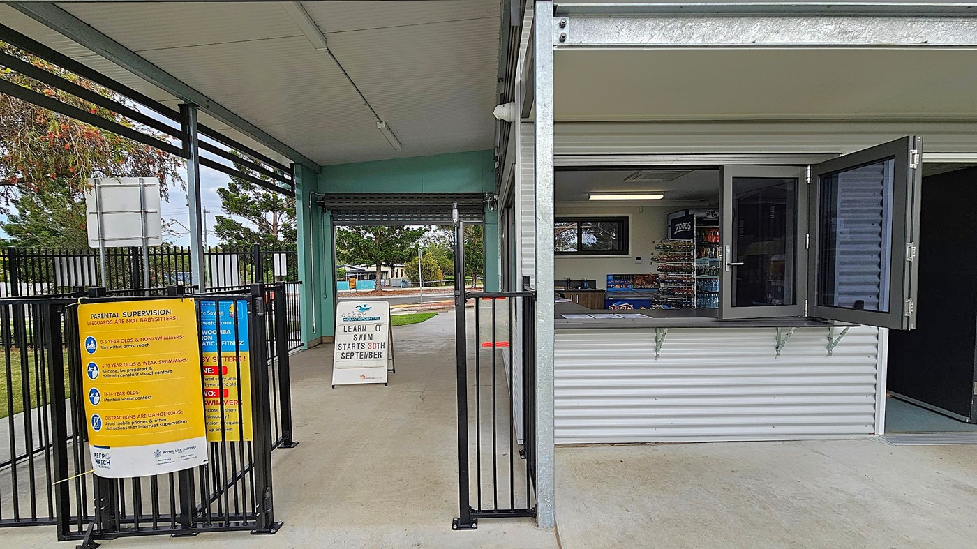 A pool entrance area with a corrugated metal building on the right, featuring an open window displaying snacks and drinks inside. A black metal gate separates the entrance from the building. On the left, a yellow sign provides parental supervision guidelines. A white sandwich board sign in the centre announces swimming lessons starting on 30th September. The floor is concrete, and the ceiling is metal. Trees and a fence are visible in the background. A pool entrance area with a corrugated metal building on the right, featuring an open window displaying snacks and drinks inside. A black metal gate separates the entrance from the building. On the left, a yellow sign provides parental supervision guidelines. A white sandwich board sign in the centre announces swimming lessons starting on 30th September. The floor is concrete, and the ceiling is metal. Trees and a fence are visible in the background.
