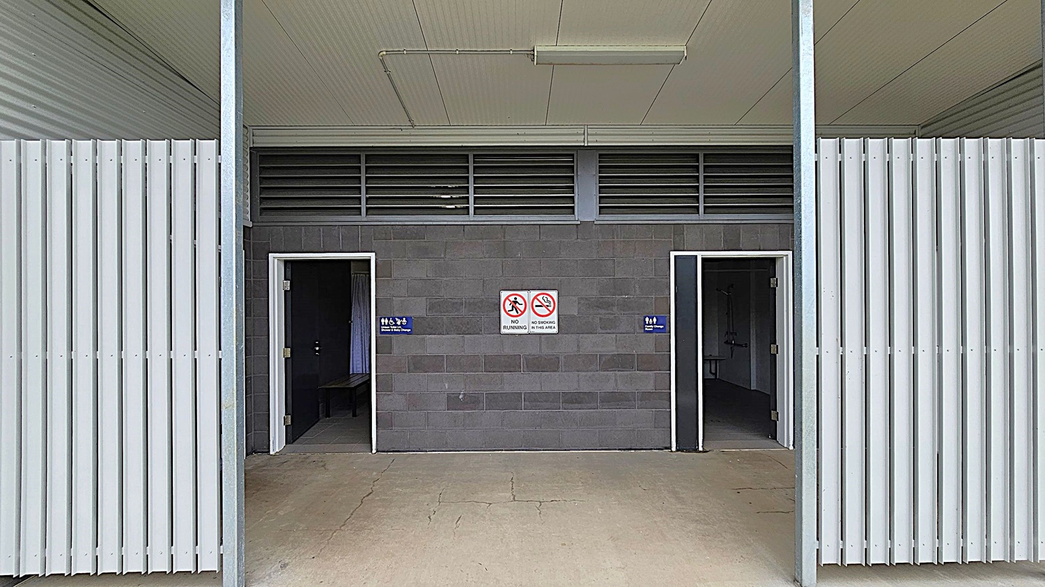 A public restroom entrance with two open doorways in a grey brick wall. The left door has a sign indicating a female toilet, and the right door indicates a male toilet. Between the doors are signs prohibiting running and smoking. The ceiling is light green with a fluorescent light fixture. The floor is concrete with visible cracks. The sides feature vertical white metal slats. A public restroom entrance with two open doorways in a grey brick wall. The left door has a sign indicating a female toilet, and the right door indicates a male toilet. Between the doors are signs prohibiting running and smoking. The ceiling is light green with a fluorescent light fixture. The floor is concrete with visible cracks. The sides feature vertical white metal slats.