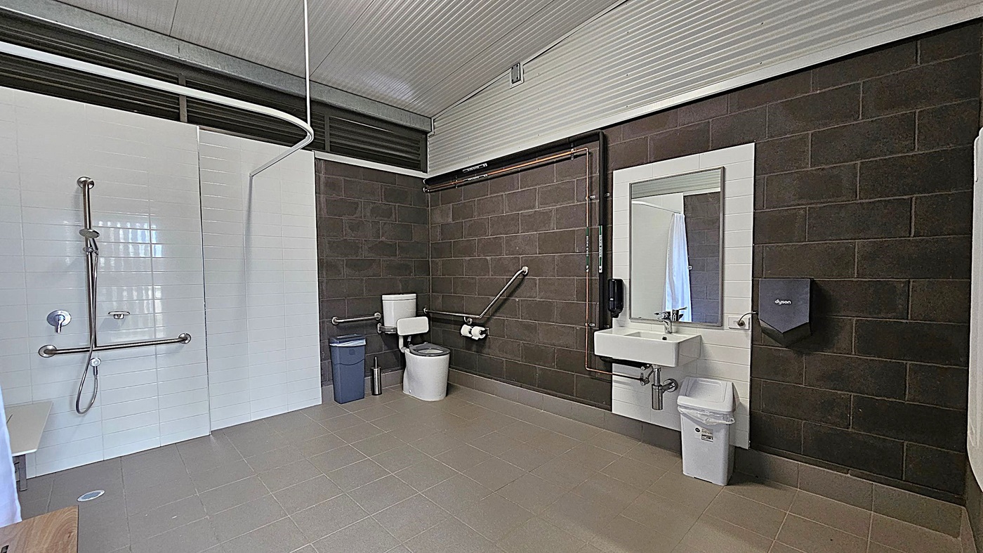 A spacious, accessible bathroom with a grey tiled floor and a corrugated metal ceiling. On the left, there is a shower area with a handheld showerhead and grab bars. In the centre, a toilet with support rails and a bin nearby. On the right, a sink with a mirror above it, a soap dispenser, and a hand dryer mounted on the dark brick wall. The walls are a mix of white tiles and dark bricks. A spacious, accessible bathroom with a grey tiled floor and a corrugated metal ceiling. On the left, there is a shower area with a handheld showerhead and grab bars. In the centre, a toilet with support rails and a bin nearby. On the right, a sink with a mirror above it, a soap dispenser, and a hand dryer mounted on the dark brick wall. The walls are a mix of white tiles and dark bricks.