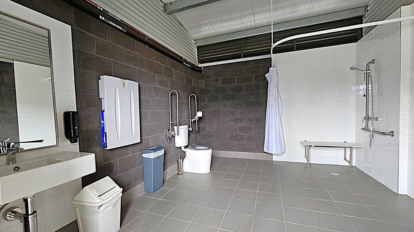 A spacious, accessible bathroom with grey tiled flooring and a corrugated metal ceiling. On the left, there's a sink with a mirror above it, a soap dispenser, and a white bin. A baby changing station is mounted on the dark grey wall. In the centre, a toilet with support rails and a blue bin nearby. On the right, a shower area with a white curtain, a bench, and grab bars. The walls are a mix of dark grey and white tiles. A spacious, accessible bathroom with grey tiled flooring and a corrugated metal ceiling. On the left, there's a sink with a mirror above it, a soap dispenser, and a white bin. A baby changing station is mounted on the dark grey wall. In the centre, a toilet with support rails and a blue bin nearby. On the right, a shower area with a white curtain, a bench, and grab bars. The walls are a mix of dark grey and white tiles.