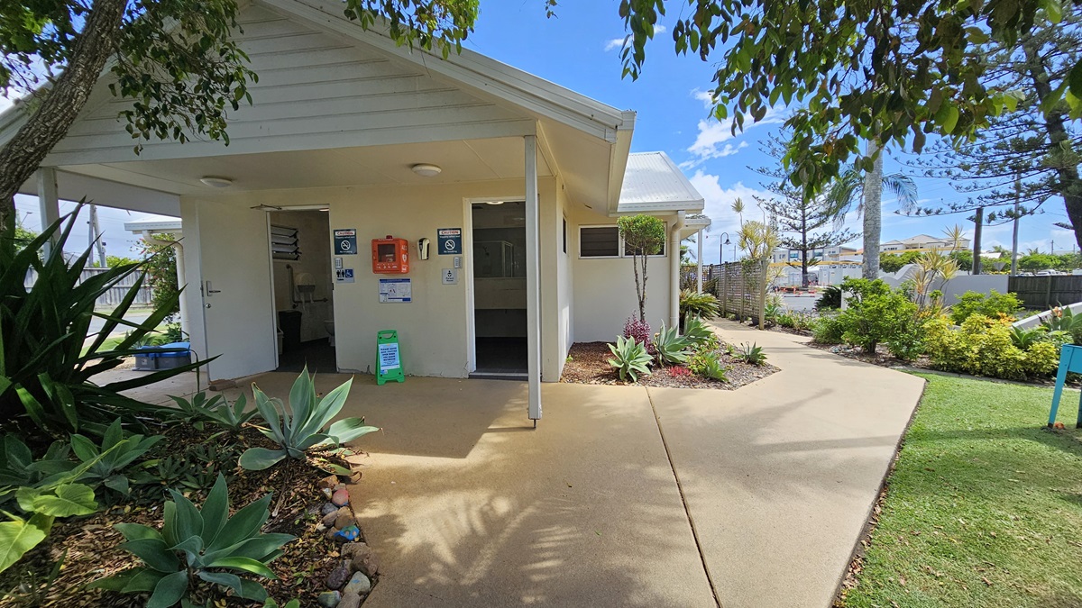A single-storey laundry building with a pitched roof and white corrugated exterior. Two manual glass doors are open, revealing the interior. Landscaped plants are positioned along the front and right side. Safety posters, notices, and a firehose reel decorate the front. A safety cone is placed nearby, while a concrete fence runs alongside the road on the right. A single-storey laundry building with a pitched roof and white corrugated exterior. Two manual glass doors are open, revealing the interior. Landscaped plants are positioned along the front and right side. Safety posters, notices, and a firehose reel decorate the front. A safety cone is placed nearby, while a concrete fence runs alongside the road on the right.