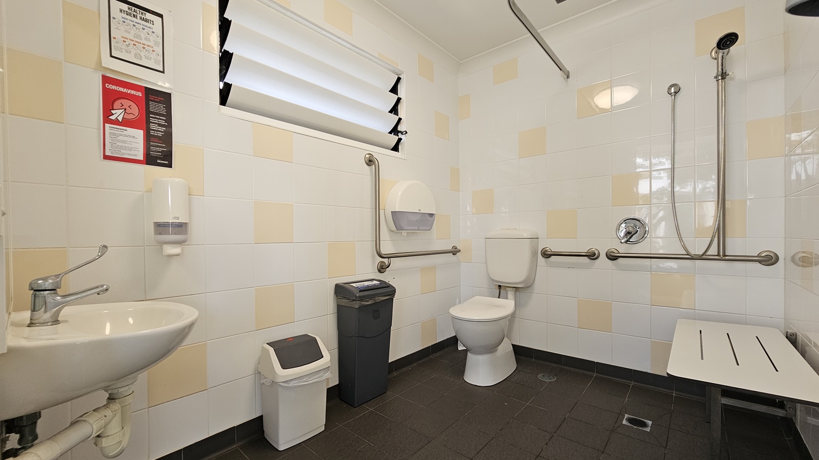 The interior of an accessible toilet. A toilet bowl with handrails is located in the corner to the left. Two rubbish bins and a tissue dispenser sit nearby. On the right wall, a shower and bench chair are mounted. A wash basin is on the left. Soap dispenser and posters adorn the wall near the window. White tiles with cream accents cover the walls, while grey tiles line the floor. The interior of an accessible toilet. A toilet bowl with handrails is located in the corner to the left. Two rubbish bins and a tissue dispenser sit nearby. On the right wall, a shower and bench chair are mounted. A wash basin is on the left. Soap dispenser and posters adorn the wall near the window. White tiles with cream accents cover the walls, while grey tiles line the floor.