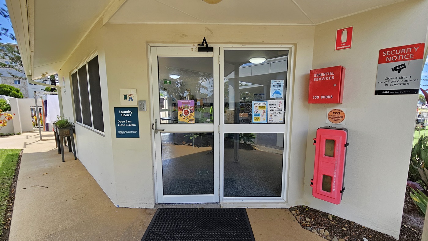 The entrance to the Laundry Room. A single-storey building with a white concrete facade. It features a manual glass sliding door with a handle. Safety stickers and posters displaying laundry hours and security reminders adorn the wall and door. A firehose reel is attached to the wall on the right. A black rubber mat lies on the doorstep. A planter box sits near the window on the left, while a landscape of plants is on the right side. The entrance to the Laundry Room. A single-storey building with a white concrete facade. It features a manual glass sliding door with a handle. Safety stickers and posters displaying laundry hours and security reminders adorn the wall and door. A firehose reel is attached to the wall on the right. A black rubber mat lies on the doorstep. A planter box sits near the window on the left, while a landscape of plants is on the right side.