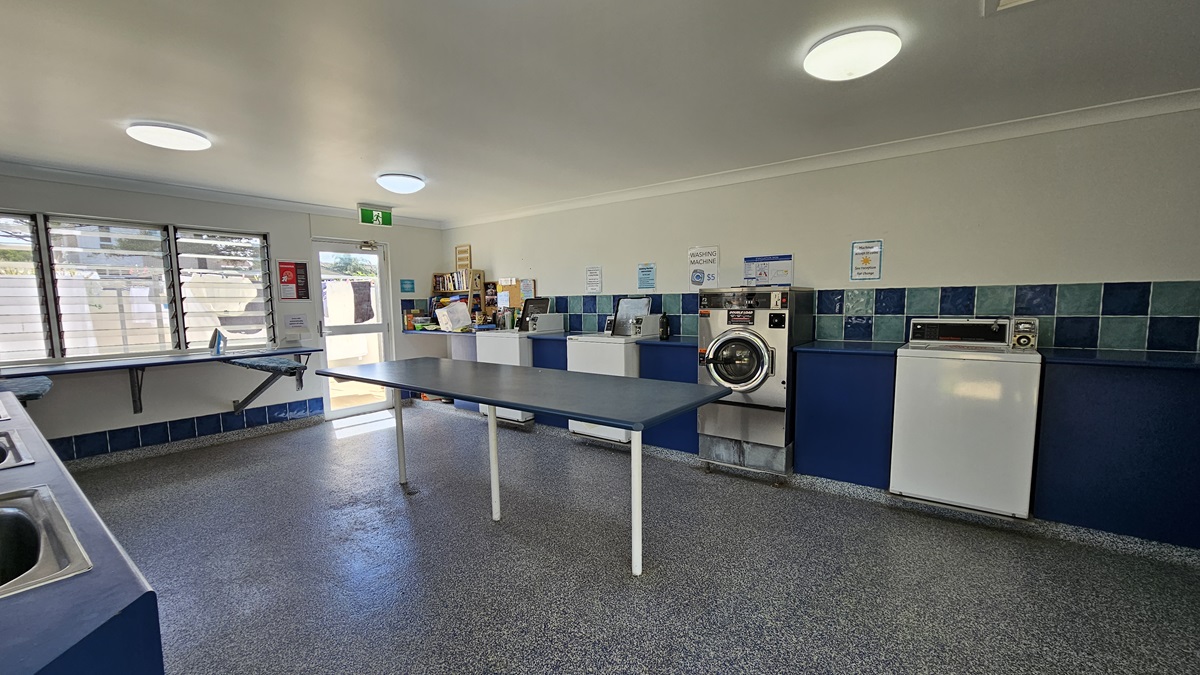 Inside the Laundry room, washing machines and dryers line the wall on the right. Three sets of sinks for hand washing are on the left, with adjacent ironing boards and irons. In the middle, a long table for folding clothes is present. Reminder posters adorn the walls. Grey tiles cover the floor, while white walls and ceiling feature LED lights. A display rack holding magazines and books is also visible. Inside the Laundry room, washing machines and dryers line the wall on the right. Three sets of sinks for hand washing are on the left, with adjacent ironing boards and irons. In the middle, a long table for folding clothes is present. Reminder posters adorn the walls. Grey tiles cover the floor, while white walls and ceiling feature LED lights. A display rack holding magazines and books is also visible.