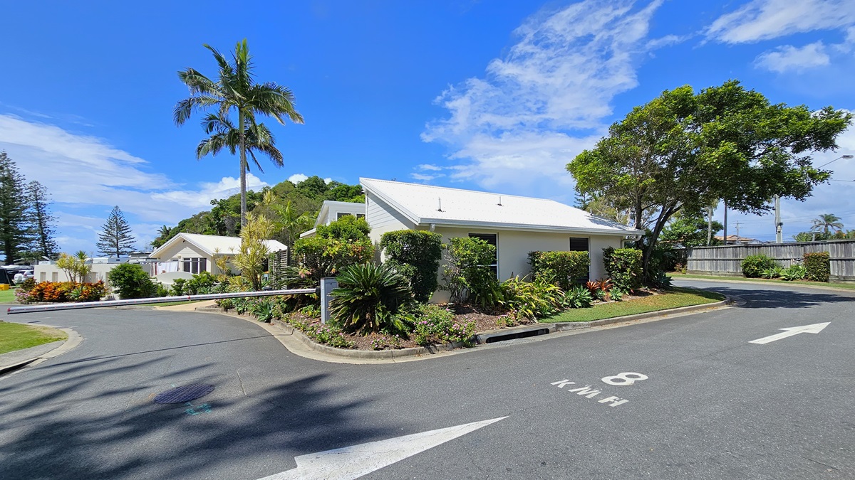 To the left, a concrete landmark displays the name "Ocean Beach Tourist Park" amid landscaped plants. The bitumen road splits, with a traffic barrier indicating no entry on the left. White markings guide travellers on the right path. Landscaped green grass fills the right side. Lamp posts and palm trees line the road. Low-set buildings with pitched roofs are positioned on the left on the left side of the road. To the left, a concrete landmark displays the name "Ocean Beach Tourist Park" amid landscaped plants. The bitumen road splits, with a traffic barrier indicating no entry on the left. White markings guide travellers on the right path. Landscaped green grass fills the right side. Lamp posts and palm trees line the road. Low-set buildings with pitched roofs are positioned on the left on the left side of the road.
