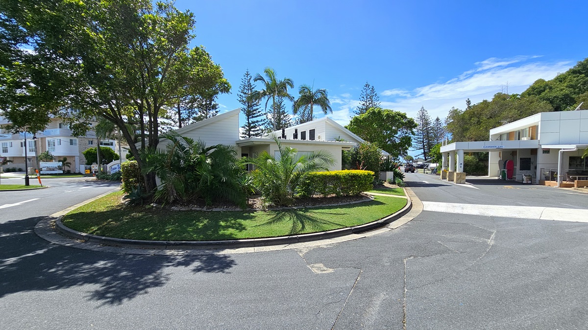 A roundabout surrounded by landscaped plants, trees, and grass. Behind it, a low-set building with a white concrete facade and pitched roof. On the right, another low-set building with a portico housing the reception office. To the left, the main road and another multi-storey building. Palm trees and pine trees are visible in the distance. A roundabout surrounded by landscaped plants, trees, and grass. Behind it, a low-set building with a white concrete facade and pitched roof. On the right, another low-set building with a portico housing the reception office. To the left, the main road and another multi-storey building. Palm trees and pine trees are visible in the distance.