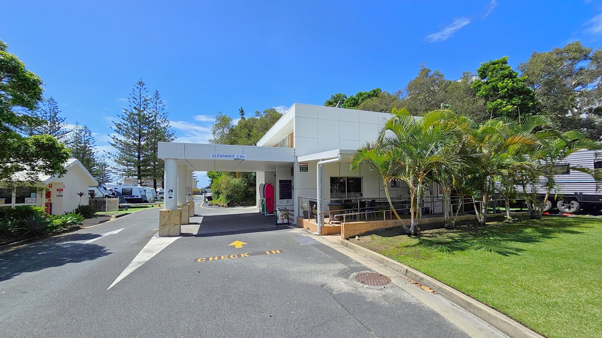 To the right is a low-set white building with a portico front. A ramp with handrails leads to the reception entrance, alongside tables and chairs. The road has markings denoting the entryway. On the left is another low-set building with a pitched roof and white concrete facade. Various trees and neatly trimmed grass are on the right. Further ahead, caravans are also visible. To the right is a low-set white building with a portico front. A ramp with handrails leads to the reception entrance, alongside tables and chairs. The road has markings denoting the entryway. On the left is another low-set building with a pitched roof and white concrete facade. Various trees and neatly trimmed grass are on the right. Further ahead, caravans are also visible.