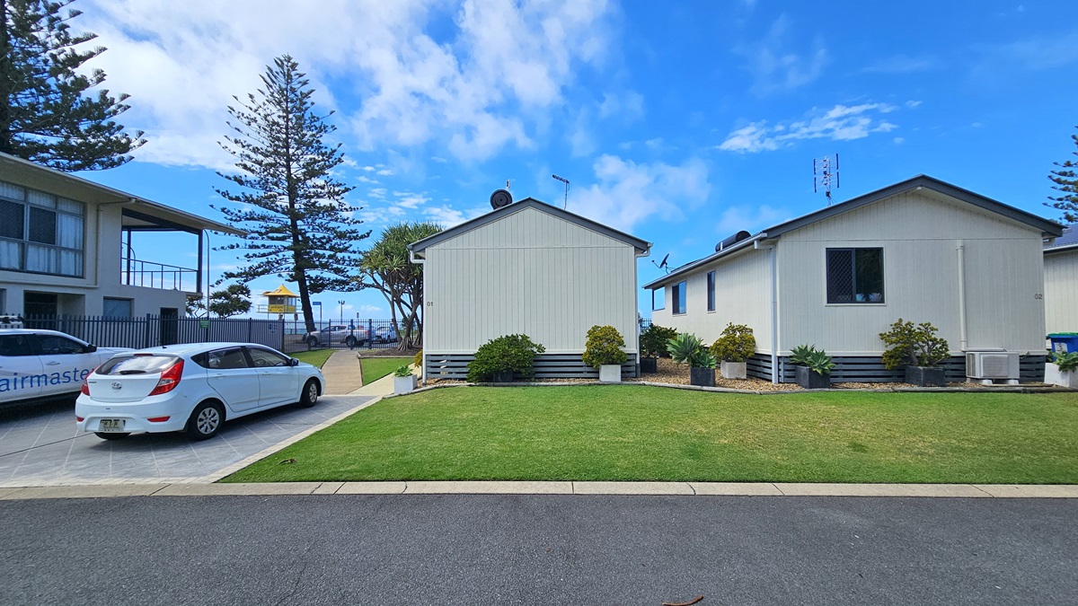 To the right, two low-set buildings with pitched roofs and white facades stand amid plants in planter boxes and green grass. To the left, a two-storey building with a veranda is accompanied by a car park with two vehicles. In the foreground, a bitumen road is visible, while in the background, the ocean and pine trees stretch out. A watchtower is also visible. To the right, two low-set buildings with pitched roofs and white facades stand amid plants in planter boxes and green grass. To the left, a two-storey building with a veranda is accompanied by a car park with two vehicles. In the foreground, a bitumen road is visible, while in the background, the ocean and pine trees stretch out. A watchtower is also visible.