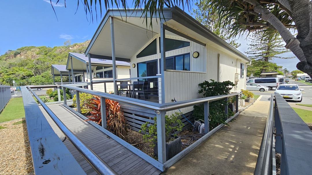 A low-set building with a white facade stands amid plants and green grass. A ramp with handrails extends from the car park to the verandah, where tables and chairs are arranged. The ramp transitions from concrete to wooden planks. Trees and grass are visible in the background. On the right is a car park occupied by a few vehicles. A low-set building with a white facade stands amid plants and green grass. A ramp with handrails extends from the car park to the verandah, where tables and chairs are arranged. The ramp transitions from concrete to wooden planks. Trees and grass are visible in the background. On the right is a car park occupied by a few vehicles.