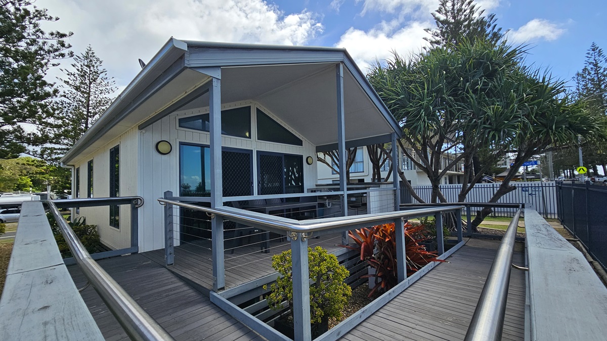 A low-set building with a white facade stands amidst plants and green grass. A ramp with handrails extends from the car park to the veranda, where tables and chairs are arranged. The ramp transitions from concrete to wooden planks. Trees and grass are visible in the background. On the right is a metal fence securing the area. A low-set building with a white facade stands amidst plants and green grass. A ramp with handrails extends from the car park to the veranda, where tables and chairs are arranged. The ramp transitions from concrete to wooden planks. Trees and grass are visible in the background. On the right is a metal fence securing the area.