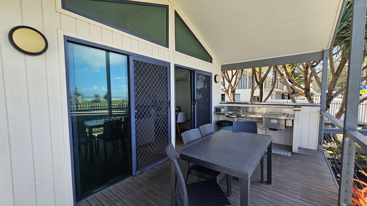The veranda features a barbecue and a sink, with movable plastic chairs and tables arranged in the middle. To the left is a glass door entrance to the cabin, equipped with a mesh door. The floor has wooden planks, and the ceiling and walls are white with light fixtures. A low fence secures the veranda, while plants and trees adorn the front. The veranda features a barbecue and a sink, with movable plastic chairs and tables arranged in the middle. To the left is a glass door entrance to the cabin, equipped with a mesh door. The floor has wooden planks, and the ceiling and walls are white with light fixtures. A low fence secures the veranda, while plants and trees adorn the front.