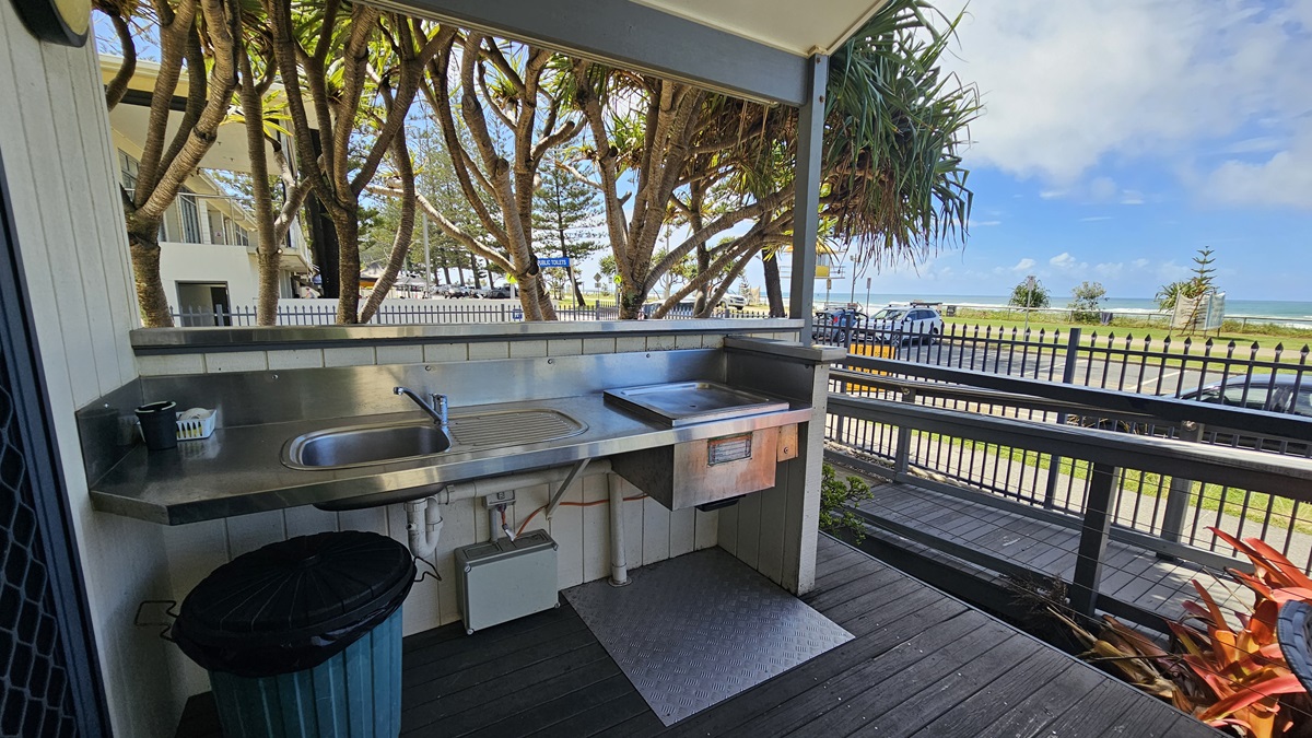 The veranda has a barbecue and a sink, with a rubbish bin placed underneath. To the left is a glass door entrance to the cabin, equipped with a mesh door. The floor has wooden planks, and the ceiling and walls are white with light fixtures. On the right, a ramp with handrails is present. A low fence secures the veranda, while plants and trees adorn the front. The veranda has a barbecue and a sink, with a rubbish bin placed underneath. To the left is a glass door entrance to the cabin, equipped with a mesh door. The floor has wooden planks, and the ceiling and walls are white with light fixtures. On the right, a ramp with handrails is present. A low fence secures the veranda, while plants and trees adorn the front.