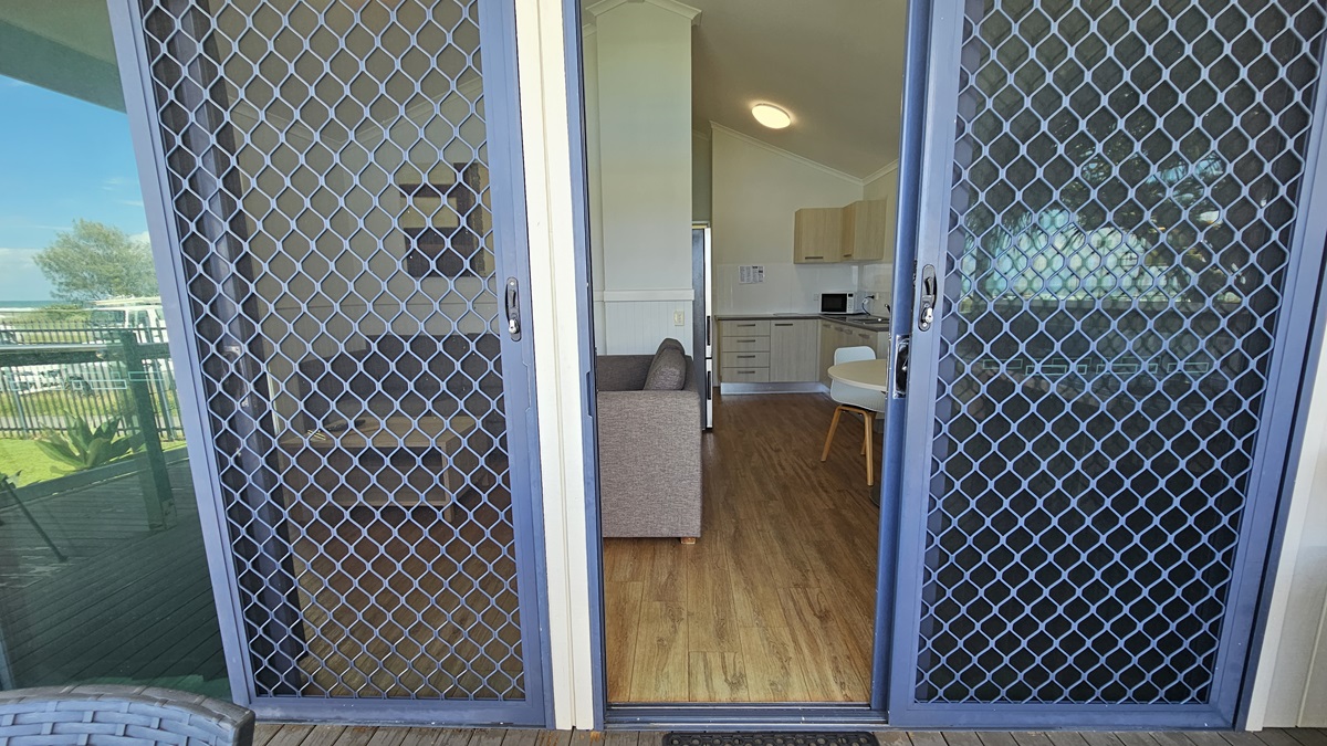 Entrance to the cabin is through a manual glass sliding door equipped with a mesh screen. The door is open, offering a view inside. The interior features a timber floor, with visible elements including the kitchen, a couch, and a dining table. Entrance to the cabin is through a manual glass sliding door equipped with a mesh screen. The door is open, offering a view inside. The interior features a timber floor, with visible elements including the kitchen, a couch, and a dining table.