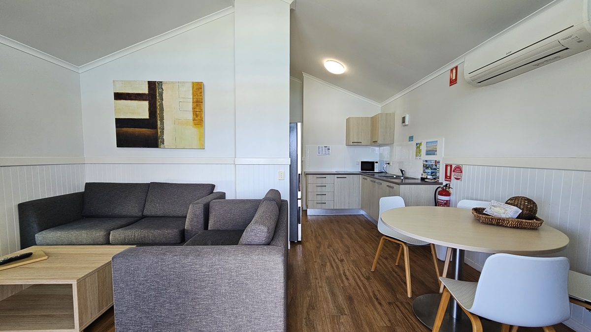 Inside the cabin, a grey couch and coffee table are set to the left, while a round table with chairs is positioned to the right. Adjacent to the table is the kitchen area, featuring a sink, kitchen cabinets, oven, and fridge. The wall is painted white with a framed decoration, and an air conditioner and fire extinguisher are mounted on it. The ceiling is white, and the floor is timber. Inside the cabin, a grey couch and coffee table are set to the left, while a round table with chairs is positioned to the right. Adjacent to the table is the kitchen area, featuring a sink, kitchen cabinets, oven, and fridge. The wall is painted white with a framed decoration, and an air conditioner and fire extinguisher are mounted on it. The ceiling is white, and the floor is timber.