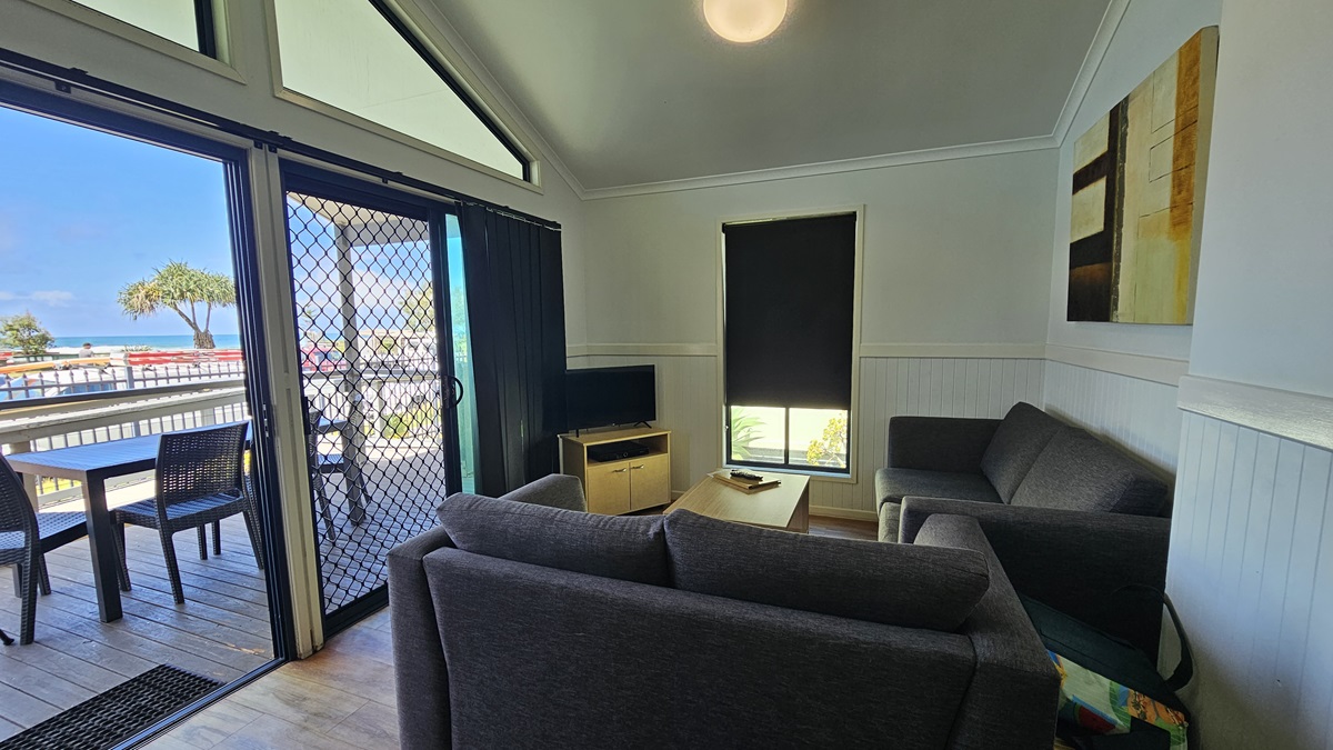 Inside the cabin, a grey couch and coffee table are positioned against the walls, facing a flatscreen TV. The wall is painted white with a framed decoration, and there's a small window in the middle. The ceiling is white with LED lights, and the floor is timber. Outside, on the veranda, there are timber floors with movable plastic tables and chairs arranged. A glass wall with a mesh screen door is on the left, along with a rubber mat. Inside the cabin, a grey couch and coffee table are positioned against the walls, facing a flatscreen TV. The wall is painted white with a framed decoration, and there's a small window in the middle. The ceiling is white with LED lights, and the floor is timber. Outside, on the veranda, there are timber floors with movable plastic tables and chairs arranged. A glass wall with a mesh screen door is on the left, along with a rubber mat.