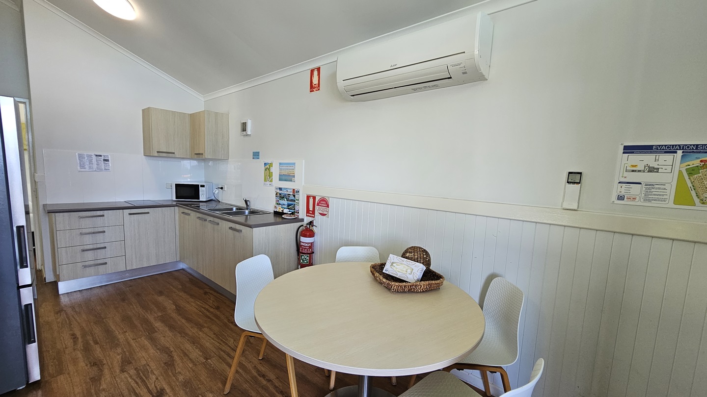 A round table with chairs is positioned to the right. Decoration and a tissue box sit on top. Adjacent to the table is the kitchen area, featuring a sink, kitchen cabinets, oven, and fridge. The wall is painted white with a poster reminder and an air conditioner and fire extinguisher are mounted on it. The pitched ceiling is white, with LED lights and the floor is timber. A round table with chairs is positioned to the right. Decoration and a tissue box sit on top. Adjacent to the table is the kitchen area, featuring a sink, kitchen cabinets, oven, and fridge. The wall is painted white with a poster reminder and an air conditioner and fire extinguisher are mounted on it. The pitched ceiling is white, with LED lights and the floor is timber.