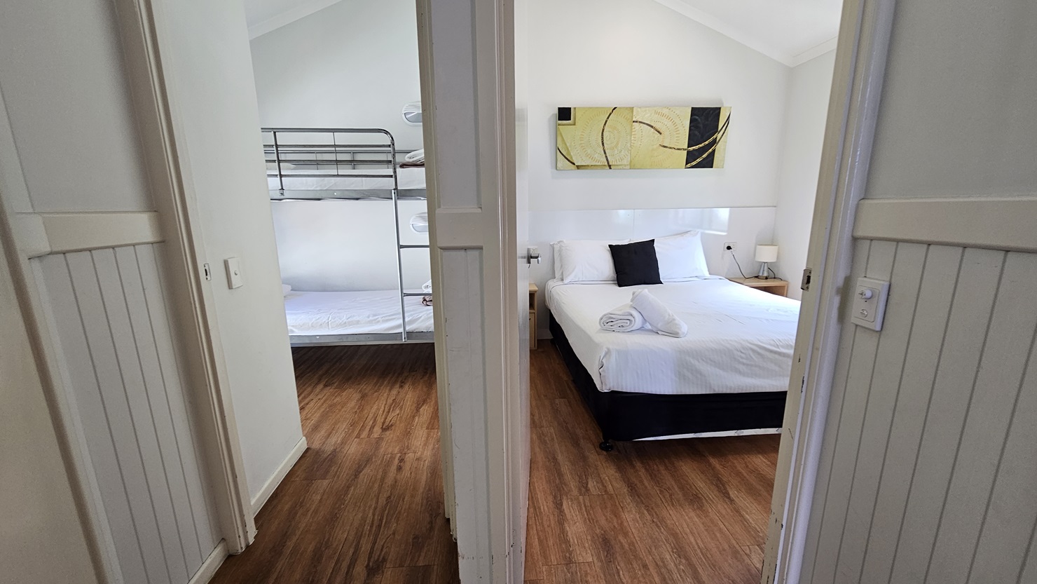 Two doors lead to the two bedrooms. The bedroom on the right features a queen bed with white covers, arranged with pillows and rolled towels. A bedside lamp sits on the right, and a framed decoration is placed above the bed. To the left is a room with a bunk bed covered in white linens. The floor is timber, and the pitched ceiling and walls are all white. Two doors lead to the two bedrooms. The bedroom on the right features a queen bed with white covers, arranged with pillows and rolled towels. A bedside lamp sits on the right, and a framed decoration is placed above the bed. To the left is a room with a bunk bed covered in white linens. The floor is timber, and the pitched ceiling and walls are all white.