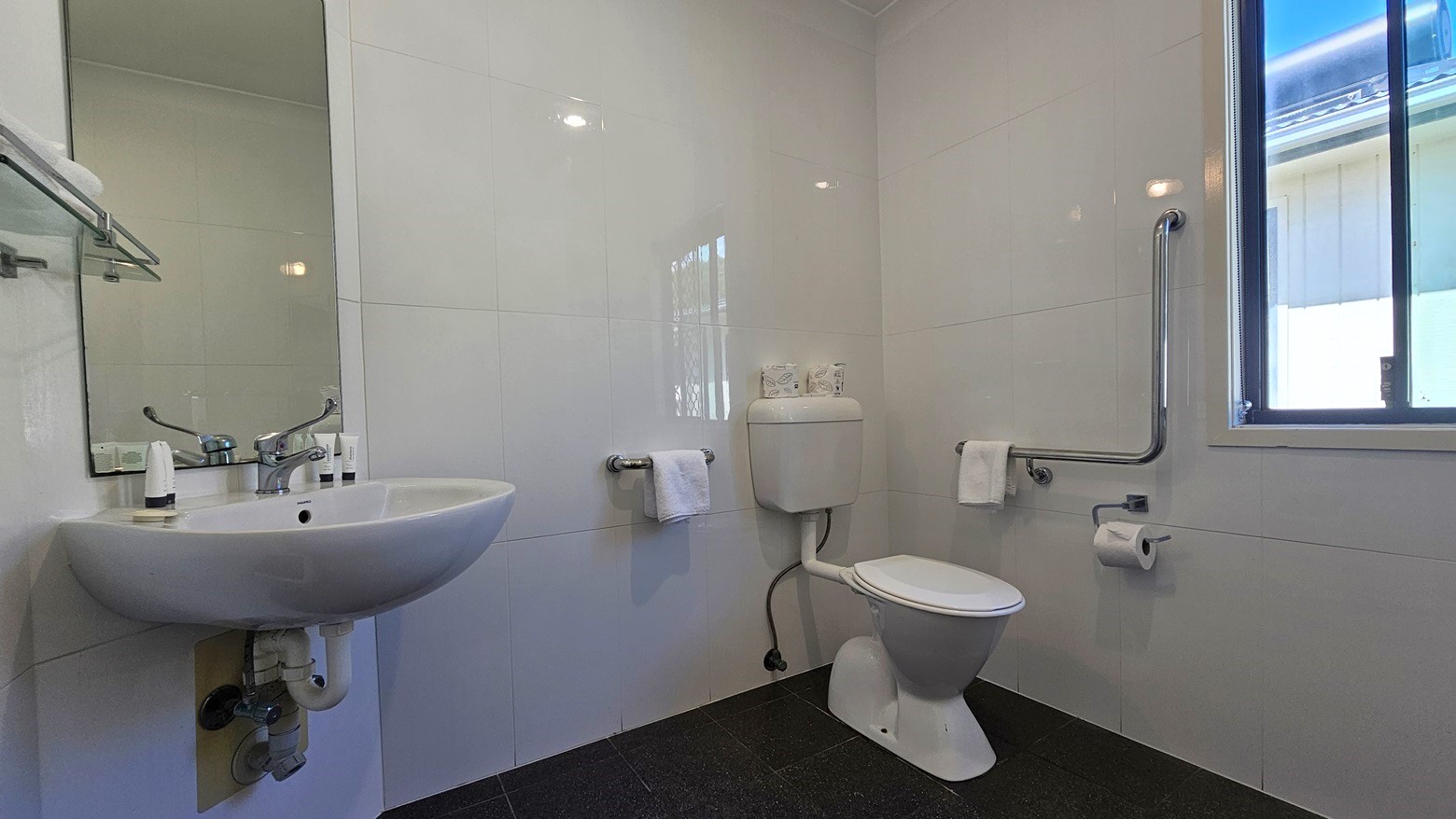 The accessible bathroom features a toilet with hand grabs positioned on the right. A glass window is on the right, adjacent to a hand basin and a wall-mounted mirror. The floor has grey tiles, and the walls are tiled with white tiles. A towel holder is mounted to the wall on the left. The accessible bathroom features a toilet with hand grabs positioned on the right. A glass window is on the right, adjacent to a hand basin and a wall-mounted mirror. The floor has grey tiles, and the walls are tiled with white tiles. A towel holder is mounted to the wall on the left.