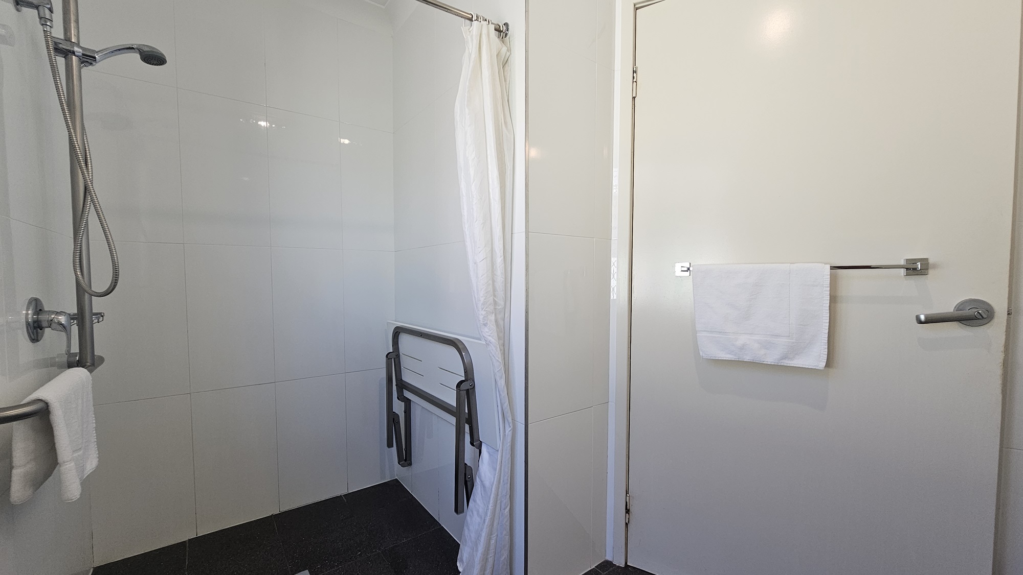 The accessible bathroom features a shower with curtains and hand grabs. A foldable bench is positioned to the right. A towel holder is mounted to the wall on the left. To the right is a door with a towel holder. The floor has grey tiles, and the walls are tiled with white tiles. The accessible bathroom features a shower with curtains and hand grabs. A foldable bench is positioned to the right. A towel holder is mounted to the wall on the left. To the right is a door with a towel holder. The floor has grey tiles, and the walls are tiled with white tiles.