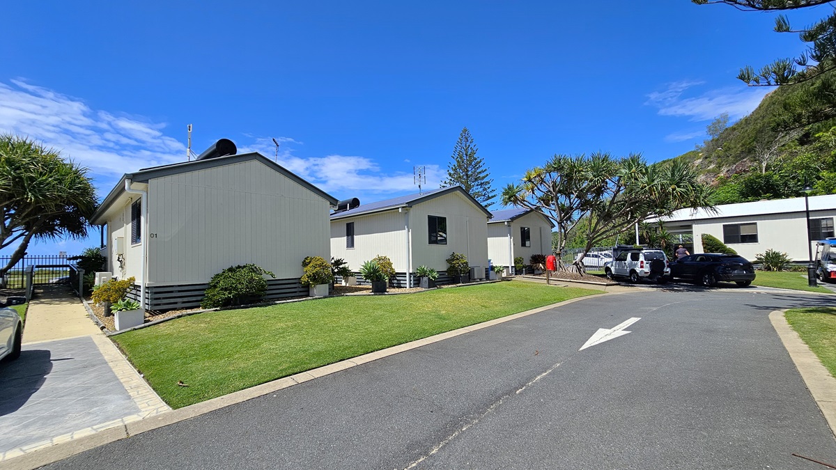 A bitumen road with markings. On the left side of the road, there is a white low set building with a dark grey roof, and windows visible on the side. In front of the building, there is a well-manicured lawn with a small garden bed hosting several plants. On the right side of the road, there is a similar white building featuring a grey roof, with its facade and entrance door visible. There are also a parked black car and a street lamp on the right side near the second building. A bitumen road with markings. On the left side of the road, there is a white low set building with a dark grey roof, and windows visible on the side. In front of the building, there is a well-manicured lawn with a small garden bed hosting several plants. On the right side of the road, there is a similar white building featuring a grey roof, with its facade and entrance door visible. There are also a parked black car and a street lamp on the right side near the second building.