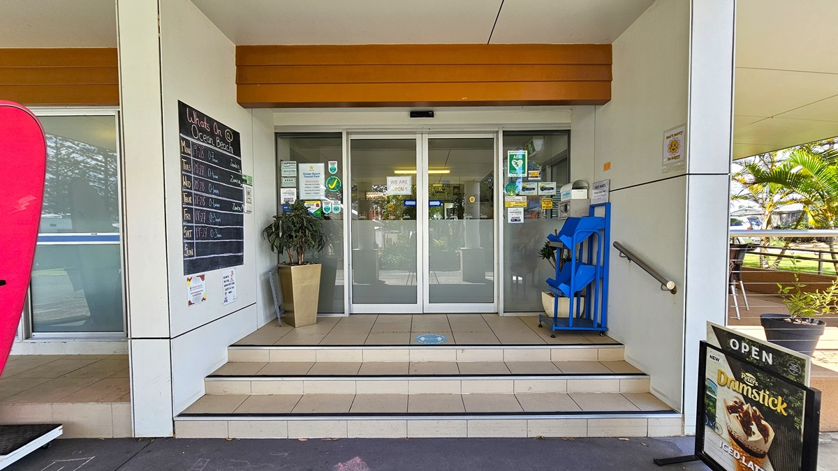 A low-set white building with a portico front. A ramp with handrails leads to the reception entrance to the right, alongside tables and chairs. On the left, a three-tiered staircase leads to an automatic sliding door entrance. Surfboards and a large poster are in front of the stairs. Trees and a truck are visible on the right. A low-set white building with a portico front. A ramp with handrails leads to the reception entrance to the right, alongside tables and chairs. On the left, a three-tiered staircase leads to an automatic sliding door entrance. Surfboards and a large poster are in front of the stairs. Trees and a truck are visible on the right.