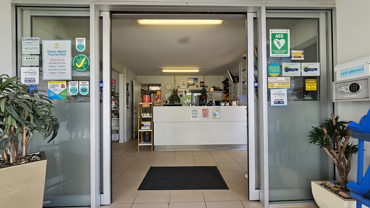In the middle is an automatic glass sliding door with a mat. Planter boxes with plants and an advertising banner are in front. Reminder posters and an access box are on the walls. Inside, a reception desk with posters and decorations beside it. The floor and ceiling are white with fluorescent lights. In the middle is an automatic glass sliding door with a mat. Planter boxes with plants and an advertising banner are in front. Reminder posters and an access box are on the walls. Inside, a reception desk with posters and decorations beside it. The floor and ceiling are white with fluorescent lights.