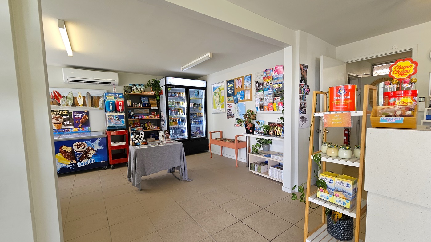 A small shop showcasing food items, drinks, and ice cream. A small table and bench chair are positioned near the wall. Posters and brochures adorn the left wall. A display rack with decorations is on the left. Cream tiles cover the floor, and the walls and ceiling are white with fluorescent lights. A small shop showcasing food items, drinks, and ice cream. A small table and bench chair are positioned near the wall. Posters and brochures adorn the left wall. A display rack with decorations is on the left. Cream tiles cover the floor, and the walls and ceiling are white with fluorescent lights.