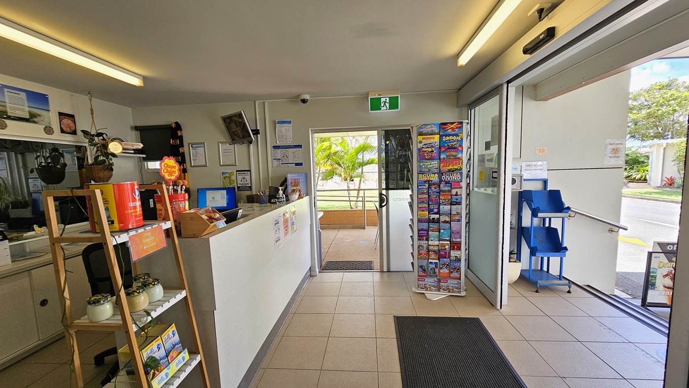 On the left, a reception desk with a computer and office accessories. Next to it, a display rack holds various items for sale. Framed posters are affixed to the wall behind the reception. A display rack filled with brochures is in the corner. Glass doors are on the right and in the middle, with a black rubber mat in front. A staircase with handrails is on the right, along with a movable rack. On the left, a reception desk with a computer and office accessories. Next to it, a display rack holds various items for sale. Framed posters are affixed to the wall behind the reception. A display rack filled with brochures is in the corner. Glass doors are on the right and in the middle, with a black rubber mat in front. A staircase with handrails is on the right, along with a movable rack.
