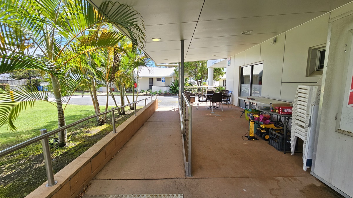 On the left is a ramp with handrails and truncated domes. Next to it, is a space with chairs and tables holding various tools. The floor is concrete, and the ceiling and walls are white. A glass door is in the middle. To the left is a grass area with palm trees. A low-set building is visible in the backdrop. On the left is a ramp with handrails and truncated domes. Next to it, is a space with chairs and tables holding various tools. The floor is concrete, and the ceiling and walls are white. A glass door is in the middle. To the left is a grass area with palm trees. A low-set building is visible in the backdrop.