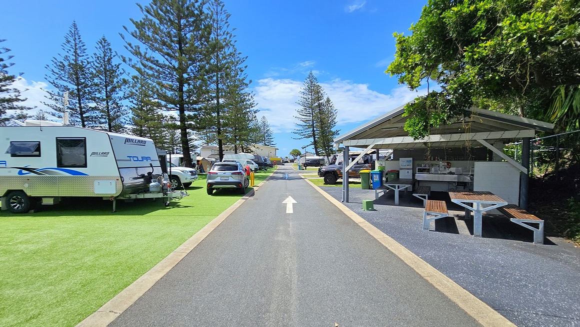 To the right is a low-set open space with a pitched roof, featuring picnic tables. On the right, barbecue, cooktop, and sink are arranged, while rubbish bins and a fridge are on the left. To the left is an area covered with synthetic turf. Rows of caravans are arranged, with a few vehicles nearby. In the middle, a road with directional arrow markings is visible. Pine trees fill the background. To the right is a low-set open space with a pitched roof, featuring picnic tables. On the right, barbecue, cooktop, and sink are arranged, while rubbish bins and a fridge are on the left. To the left is an area covered with synthetic turf. Rows of caravans are arranged, with a few vehicles nearby. In the middle, a road with directional arrow markings is visible. Pine trees fill the background.