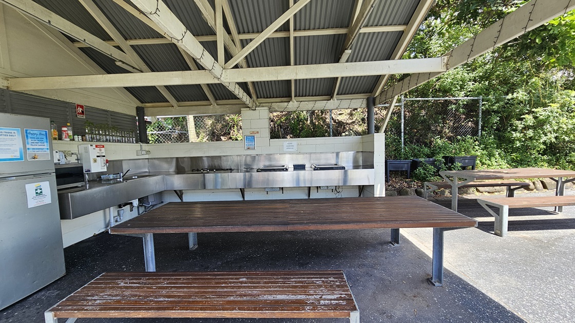 The barbecue area comprises a low-set open building with a pitched corrugated roof and exposed metal beams. Wooden picnic tables are arranged underneath. Barbecues, cooktops, and a sink with kitchen accessories and condiments are against the wall. A fridge stands on the left. A chain fence secures the area, while wild grass and planter boxes are visible behind another picnic table on the right. The barbecue area comprises a low-set open building with a pitched corrugated roof and exposed metal beams. Wooden picnic tables are arranged underneath. Barbecues, cooktops, and a sink with kitchen accessories and condiments are against the wall. A fridge stands on the left. A chain fence secures the area, while wild grass and planter boxes are visible behind another picnic table on the right.