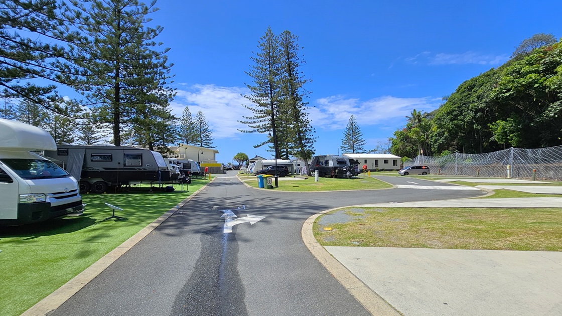 On the left, there are several caravans parked on artificial turfs with vehicles attached to the front of some caravans. Trees line the background, On the right, there is a paved road with a footpath adjacent to it. More caravans are parked on the grass further along the right side. Tall pine trees stand prominently in the centre of the image separating the left and right sections. A low-set building is also visible on the far right. The chain fence secures the right. On the left, there are several caravans parked on artificial turfs with vehicles attached to the front of some caravans. Trees line the background, On the right, there is a paved road with a footpath adjacent to it. More caravans are parked on the grass further along the right side. Tall pine trees stand prominently in the centre of the image separating the left and right sections. A low-set building is also visible on the far right. The chain fence secures the right.