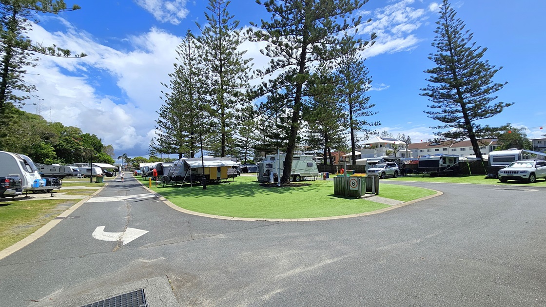 Several caravans are parked on artificial turfs on the left, with vehicles attached to some. Trees line the background. On the right, a paved road is visible, with more caravans parked on grass further along. Tall pine trees stand prominently in the centre, with additional caravans separating the left and right sections. Buildings and more pine trees are visible on the far right. Several caravans are parked on artificial turfs on the left, with vehicles attached to some. Trees line the background. On the right, a paved road is visible, with more caravans parked on grass further along. Tall pine trees stand prominently in the centre, with additional caravans separating the left and right sections. Buildings and more pine trees are visible on the far right.