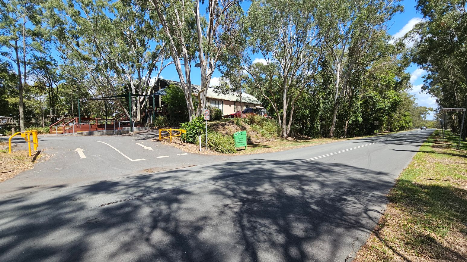 A road stretches into the distance on the right, bordered by tall trees casting shadows. On the left, a driveway leads to a building partially obscured by trees, with a small green sign and yellow barriers at the entrance. The ground is covered with fallen leaves, and the sky is clear with a few clouds.