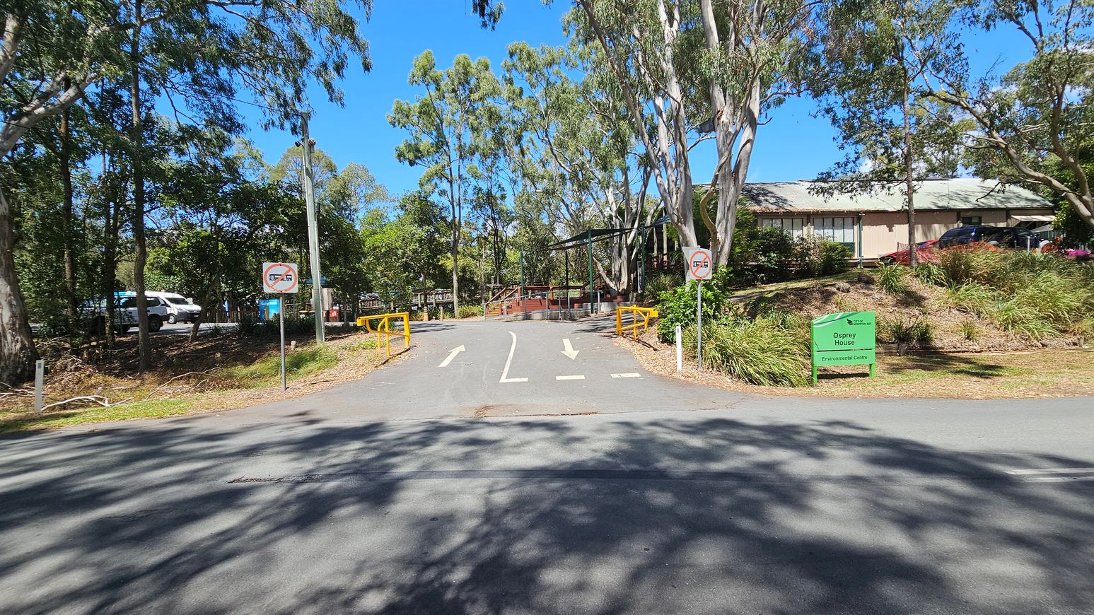 A paved road leads to a building partially visible on the right, surrounded by trees and greenery. Yellow barriers and directional arrows are on the road. Two signs indicate no U-turns. A green sign on the right reads "Osprey House Environmental Centre." On the left, a parked white vehicle is visible among the trees. The area is shaded by tall trees, and the sky is clear and blue.