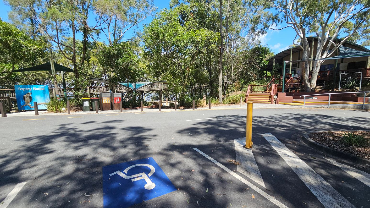 A parking area with a blue disabled parking space marked on the asphalt in the foreground. A yellow bollard is positioned near the centre. In the background, there are trees and a building with a wooden deck and steps. To the left, a sign reads "Osprey House" with an image of an osprey. Several bins are lined up near the sign. The area is surrounded by greenery, with shadows of trees cast on the ground.