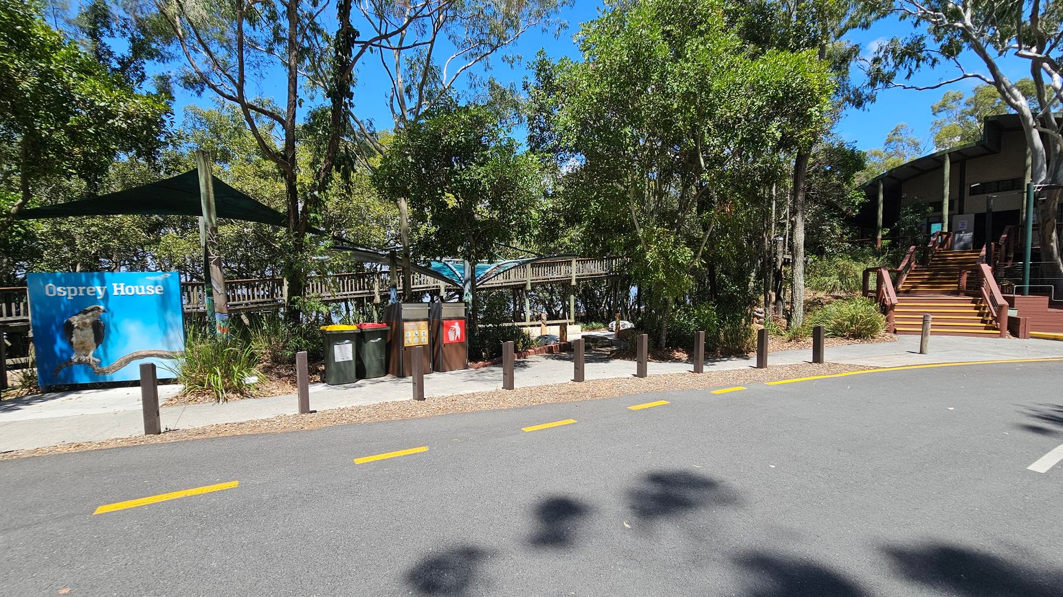 A paved road with yellow markings runs across the foreground. On the left, a sign reads "Osprey House" with an image of a bird. Nearby, several bins are placed under a shaded area. In the centre, a wooden walkway is partially visible, surrounded by dense trees. On the right, a building with wooden steps leads up to an entrance, partially obscured by trees. The area is lush with greenery, and the sky is clear and blue.