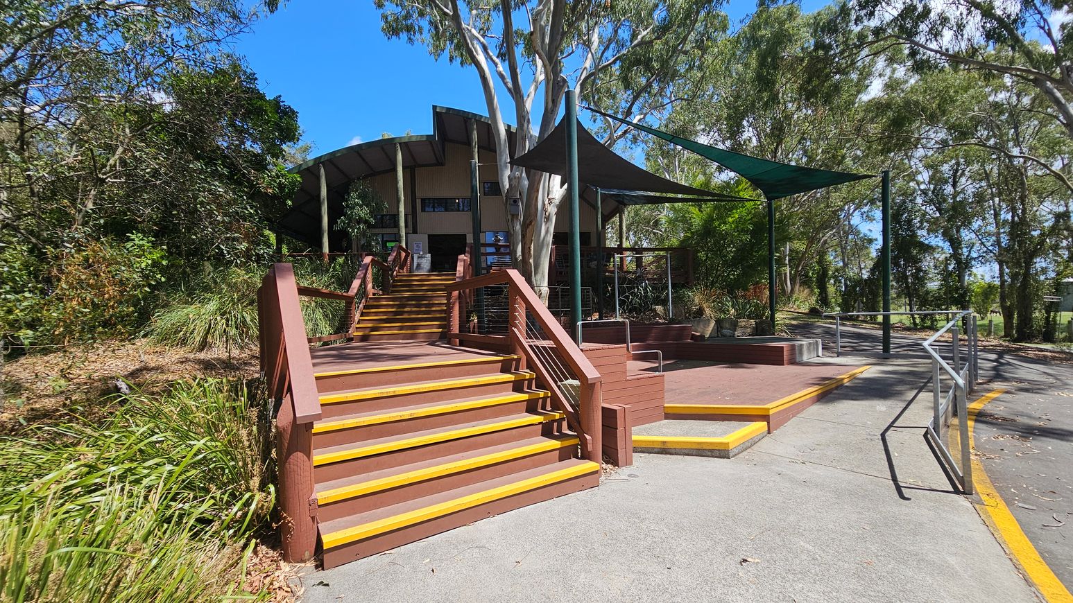 A set of brown stairs with yellow edges leads up to a building with a curved roof. The building is partially obscured by tall trees. On the right, there is a paved path with a metal railing and a shaded seating area with a green canopy. The left side is lined with dense greenery and bushes. The ground is a mix of concrete and brown decking.