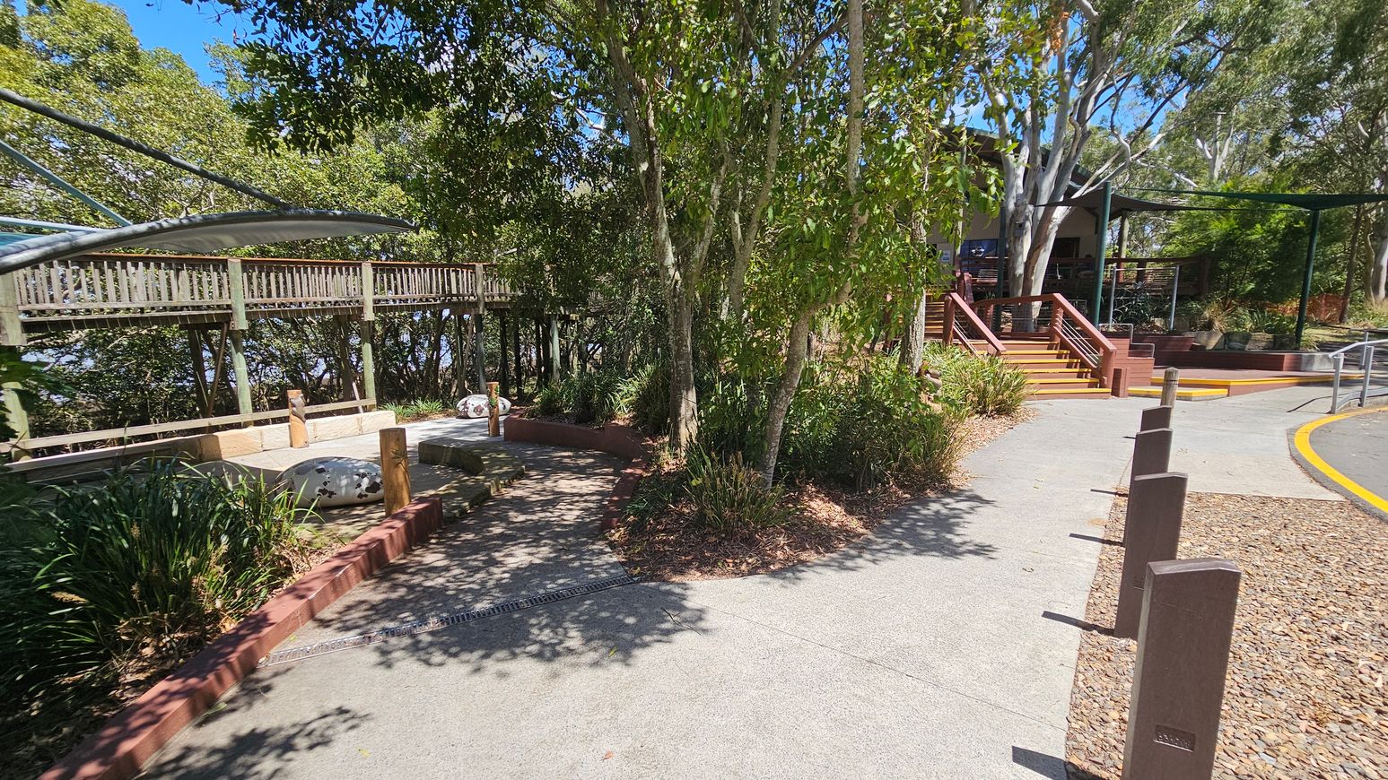 A pathway curves through a park area with trees and greenery on both sides. On the left, there's a wooden walkway elevated on stilts, partially covered by a curved metal canopy. In the centre, a small seating area with benches and decorative elements is surrounded by plants. On the right, a set of wooden steps leads up to a building with a shaded area. The ground is a mix of concrete and gravel, with wooden posts lining the path.