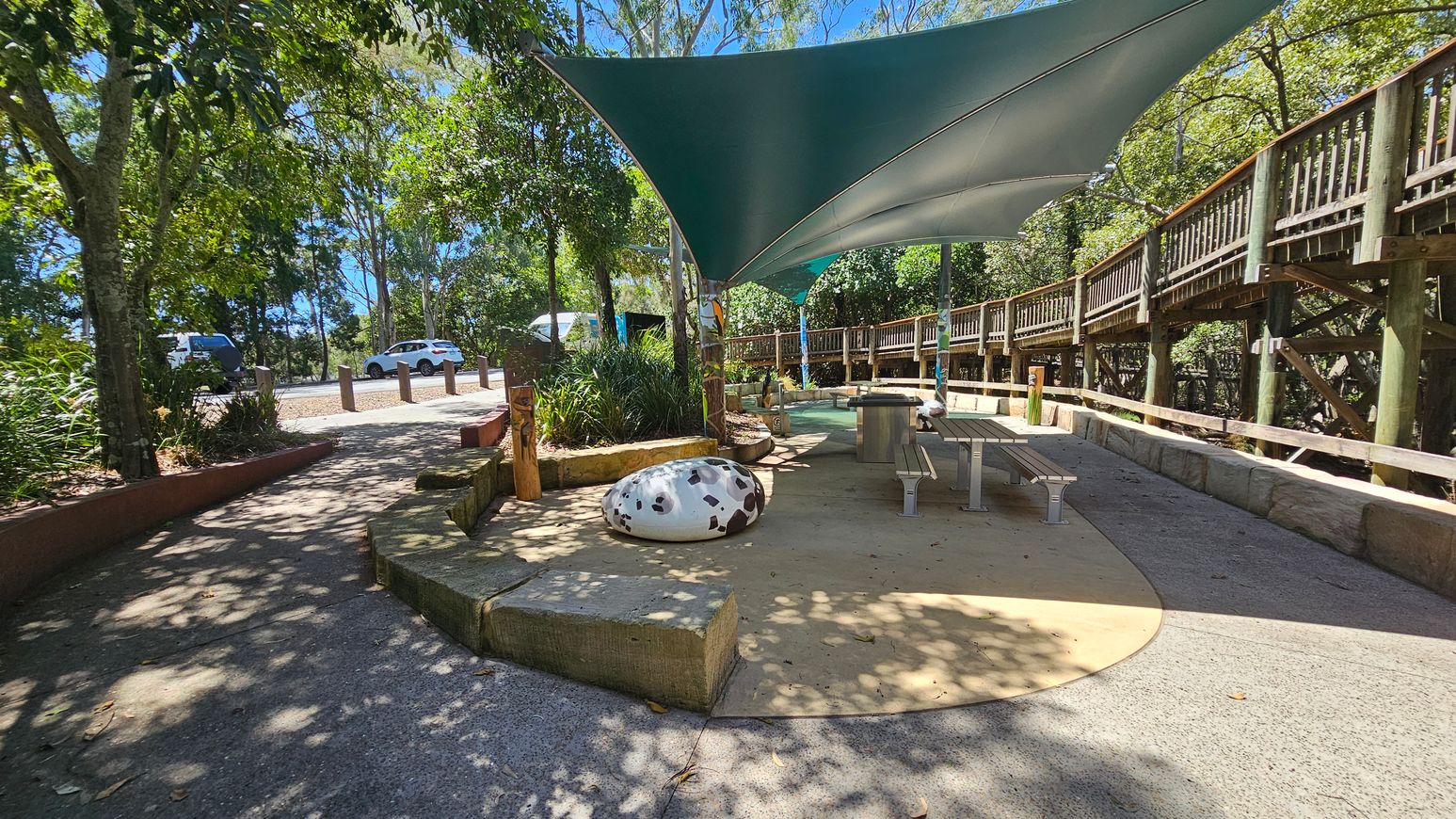 A shaded outdoor area with a green canopy overhead. On the right, there is a wooden walkway with railings elevated above the ground. Below the canopy, there are picnic tables and a large, spotted decorative stone. The ground is paved with a mix of concrete and stone. To the left, trees and shrubs provide natural shade, and a few cars are parked on a gravel path. The area is surrounded by lush greenery.