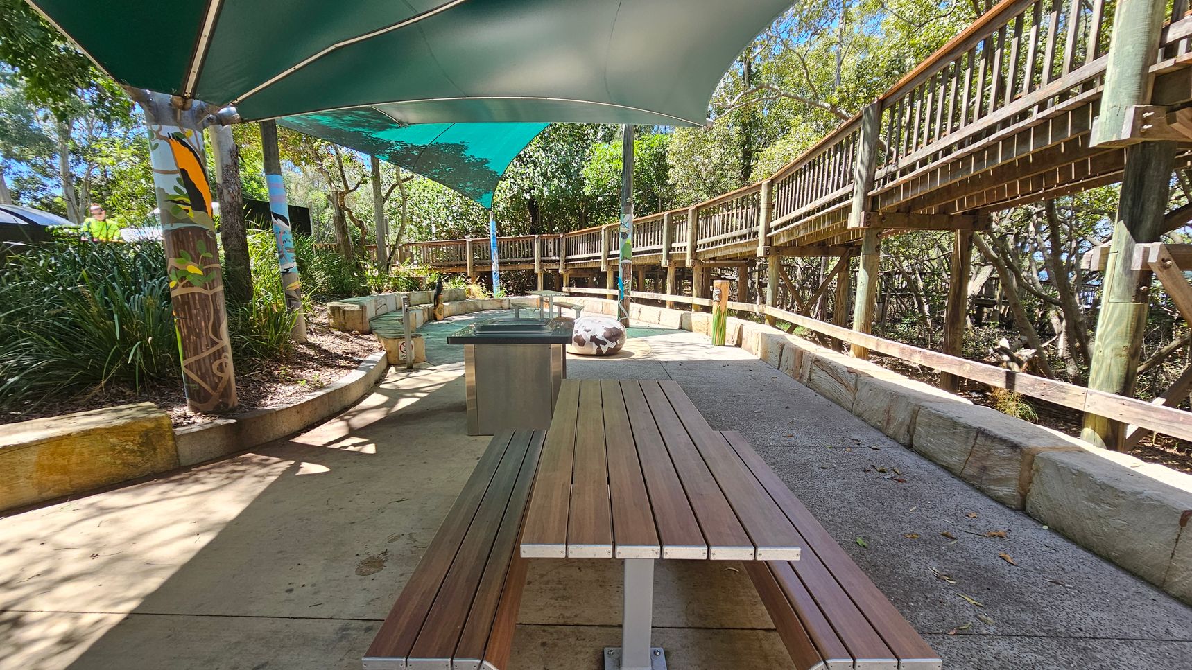 A shaded outdoor area with a wooden picnic table in the foreground. The ground is paved with concrete. To the right, a wooden walkway with railings runs parallel to the area, elevated on posts. In the background, there are trees and greenery, providing a natural setting. The ceiling consists of green fabric canopies supported by poles, some of which are painted with colourful designs. A stone bench and a round stone feature are visible in the middle distance.