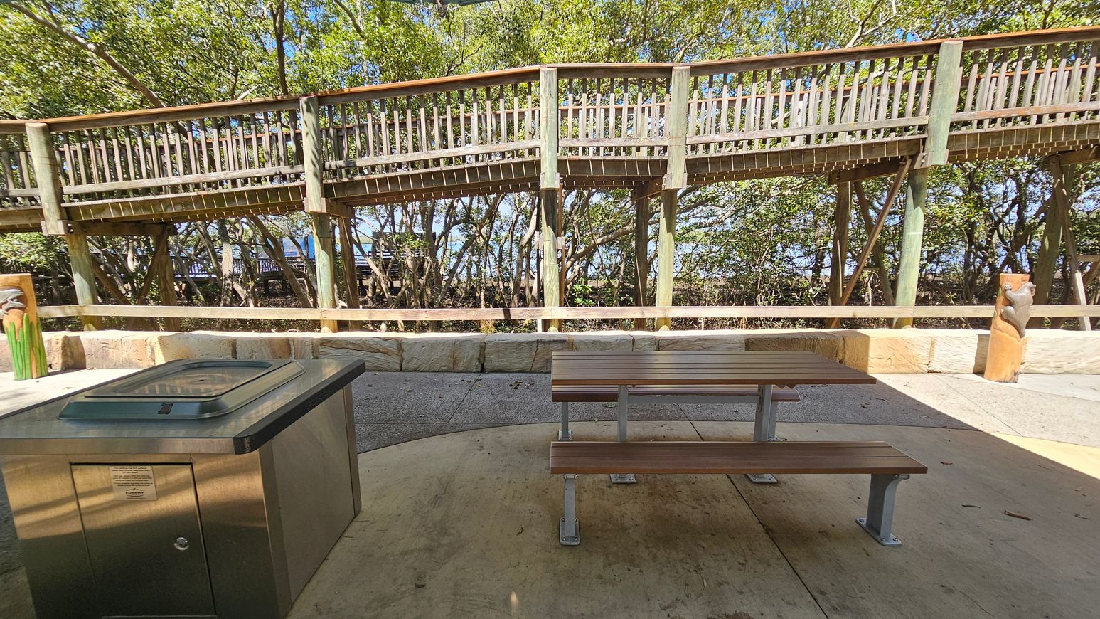 A wooden picnic table with metal legs is positioned on a concrete surface in the foreground. To the left, there is a stainless steel barbecue grill. In the background, a wooden boardwalk supported by posts runs horizontally, with trees and foliage visible behind it. The ground is a mix of concrete and stone, and there is a low stone wall separating the picnic area from the boardwalk.