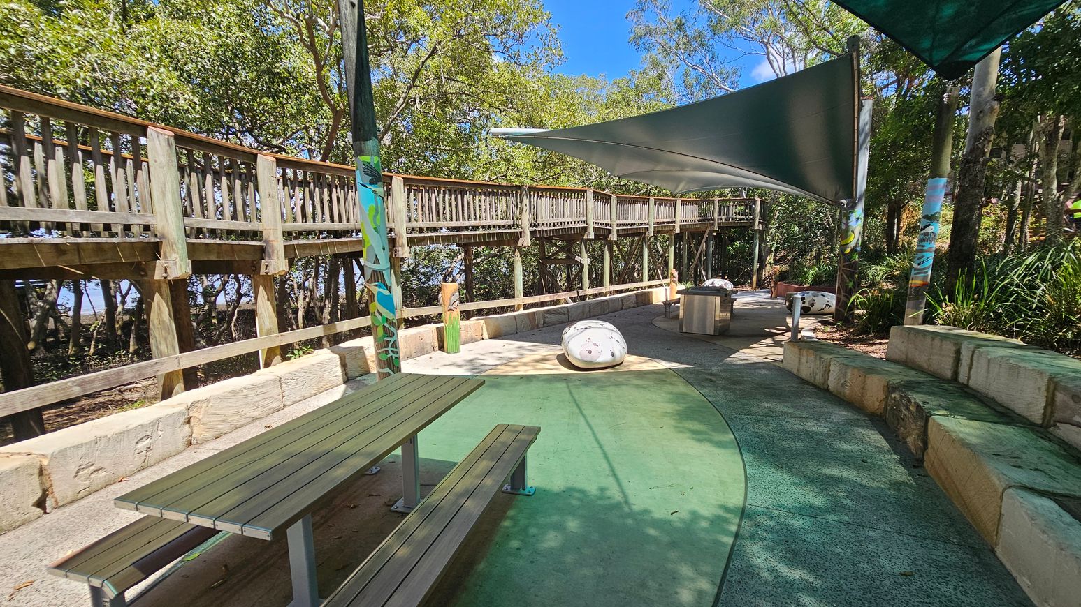 A wooden boardwalk runs along the left side, elevated and supported by posts, with trees visible behind it. In the centre, there's a shaded area with a green canopy, a picnic table, and a large, smooth stone. The ground is a mix of green and concrete surfaces. On the right, there are stone benches and more greenery. The area is surrounded by trees, providing a natural setting.