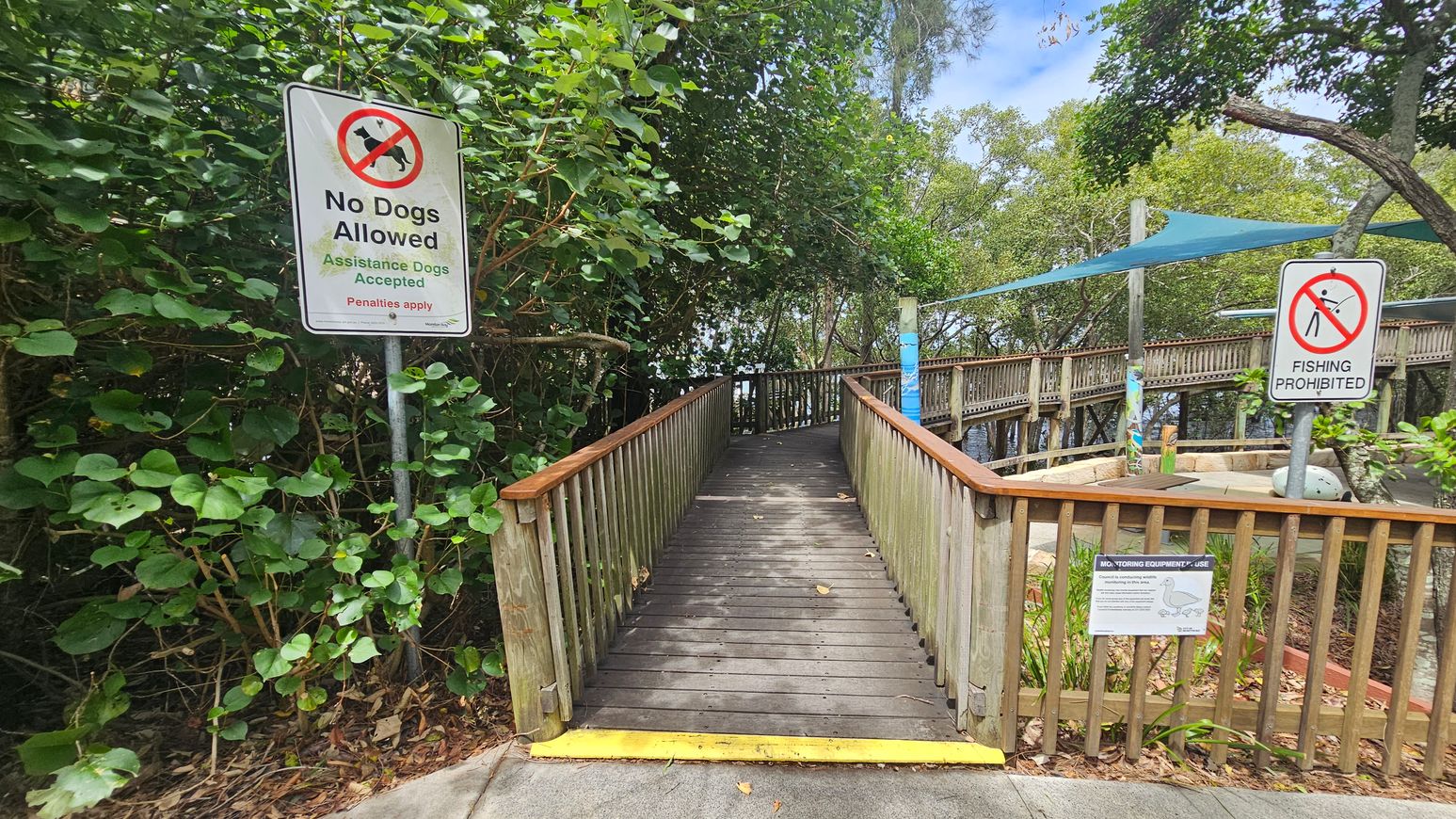 A wooden walkway with railings leads into a lush, green area. On the left, a sign reads "No Dogs Allowed" with an exception for assistance dogs. On the right, another sign states "Fishing Prohibited." The walkway is slightly elevated, with a yellow strip at the entrance. Surrounding the path are dense trees and foliage, providing shade. A blue shade cloth is visible in the background, covering part of the area. The ground is covered with leaves and the path is made of wooden planks.