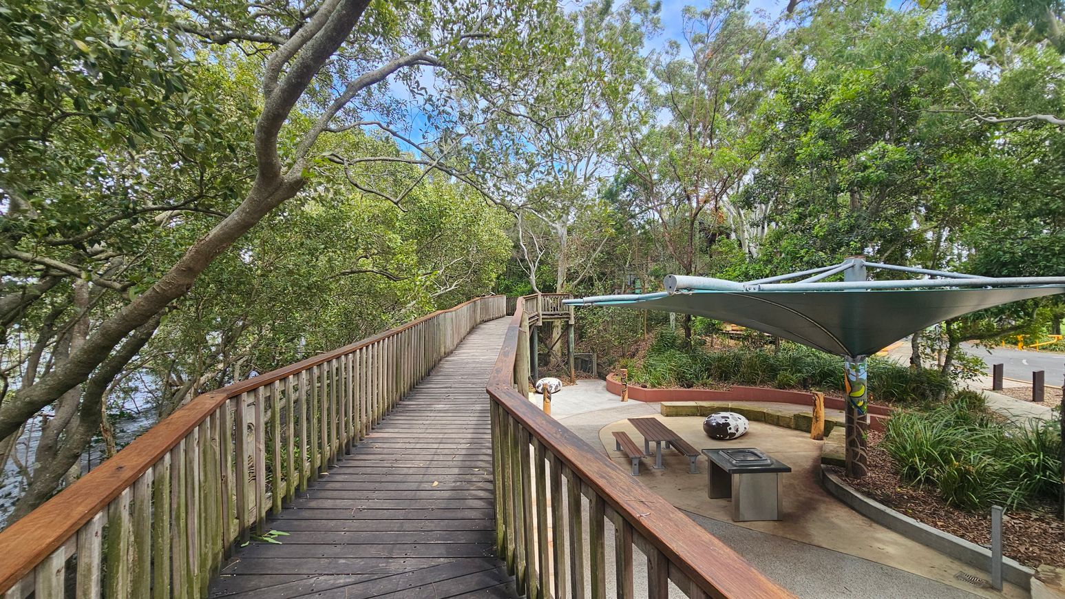 A wooden boardwalk with a railing on the left curves through a lush, green forest area. On the right, there's a small seating area with benches and a table under a large, modern canopy. The ground is paved, and there are plants and trees surrounding the area. A path leads away from the seating area, and a road is visible in the background. The scene is shaded by the dense tree canopy above.