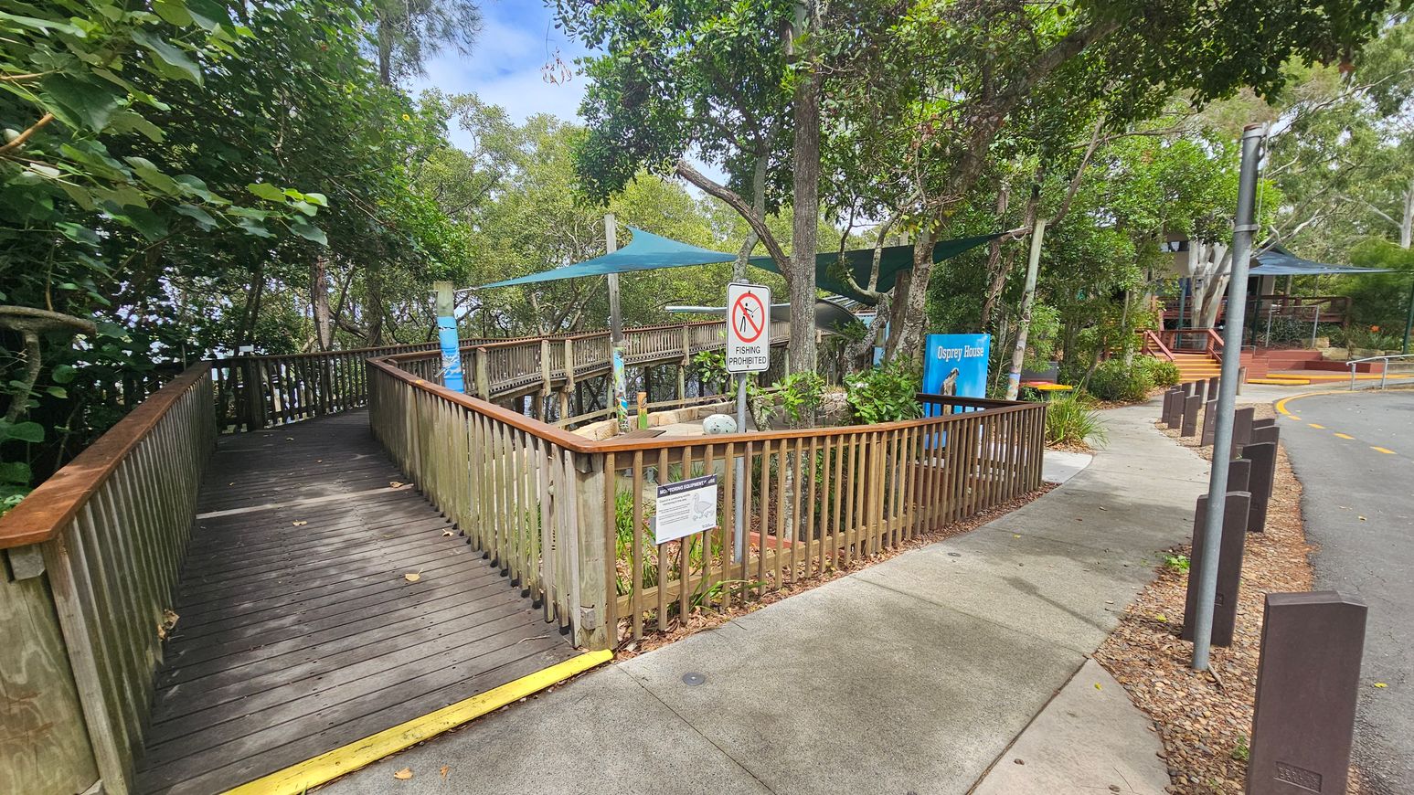 A wooden boardwalk on the left leads into a wooded area with dense green foliage. A sign on the railing reads "Fishing Prohibited." In the centre, a blue sign indicates "Osprey House." The right side features a paved path with bollards and a building with a shaded area. The ground is a mix of concrete and gravel, and the area is surrounded by trees.