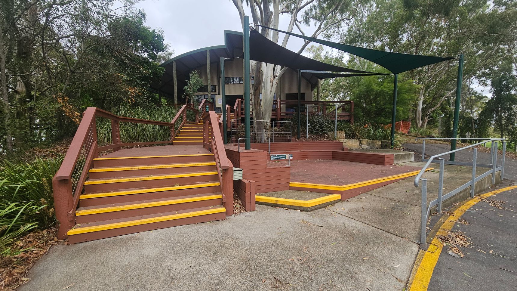 A pathway leads to a building with a curved roof in the background. In the foreground, there are wide steps with yellow edges and wooden railings on the left. A ramp with metal railings is on the right. The area is surrounded by trees and greenery, with a large tree near the building. A shade sail is stretched above the entrance area. The ground is a mix of concrete and red paving, with some leaves scattered around.